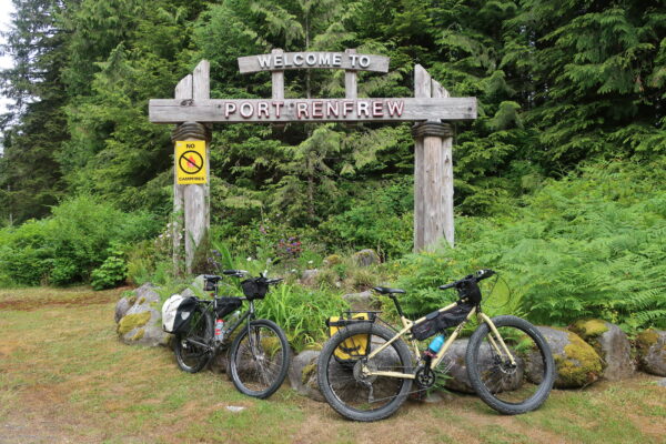 Two bicyclists in front of Welcome to Port Renfrew sign on Vancouver Island British Columbia